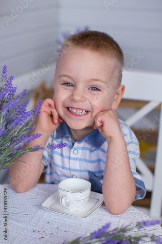 A child boy sits at a table in the scenery of Provence. White table and chair with bouquets of lavender on the background of the facade with shutters. On the table is a white cup and saucer