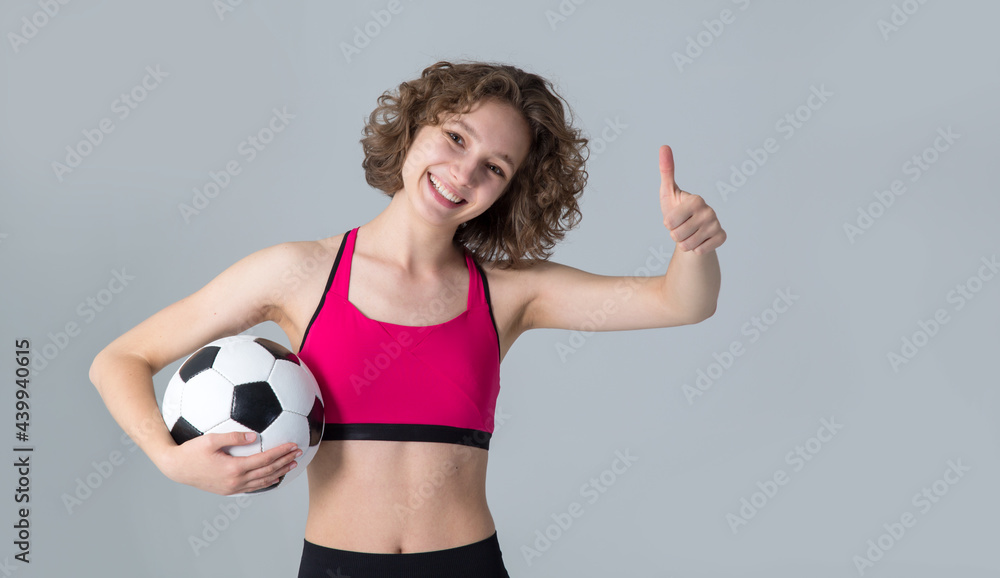 Sportive woman with soccer ball shows thumb up, standing against gray background. Sportive woman with soccer ball shows thumb up, standing against gray background.
