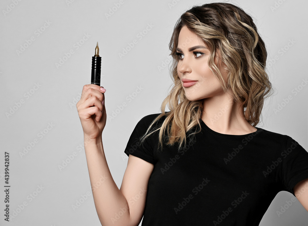 Portrait of young curly blonde woman, permanent makeup master in black t-shirt with copy space holding eyebrow tattooing professional machine, equipment in hand over grey background Portrait of young curly blonde woman, permanent makeup master in black t-shirt with copy space holding eyebrow tattooing professional machine, equipment in hand over grey background