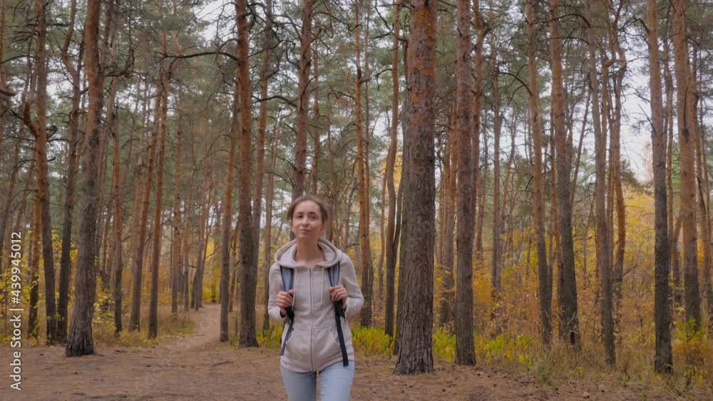 Front view of young positive woman in hoodie with backpack walking in autumn park, coniferous forest - wide angle steadicam shot. Active outdoor lifestyle, leisure time, freedom and adventure concept Front view of young positive woman in hoodie with backpack walking in autumn park, coniferous forest - wide angle steadicam shot. Active outdoor lifestyle, leisure time, freedom and adventure concept