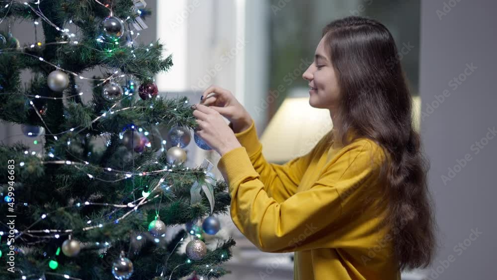 Side view of young charming woman making a wish hanging decoration on New Year tree. Medium shot portrait of beautiful slim Middle Eastern lady with long brunette hair decorating home on Christmas eve Side view of young charming woman making a wish hanging decoration on New Year tree. Medium shot portrait of beautiful slim Middle Eastern lady with long brunette hair decorating home on Christmas eve