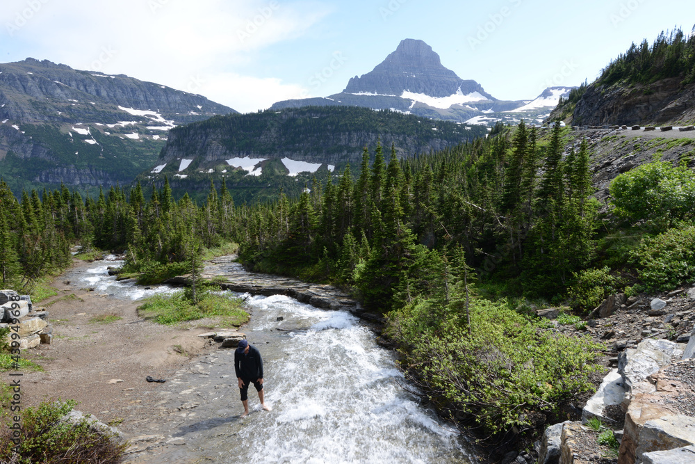 A man taking a closer look at a river in Glacier National Park, with snow capped mountains in the background