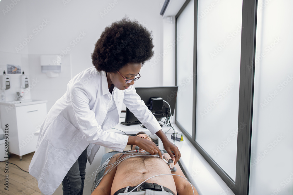 © sofiko14 - Medicine, health and heart check-up concept. Young afro-american woman doctor with electrocardiogram equipment making cardiogram test to male patient in hospital, fixing electrodes on patient chest