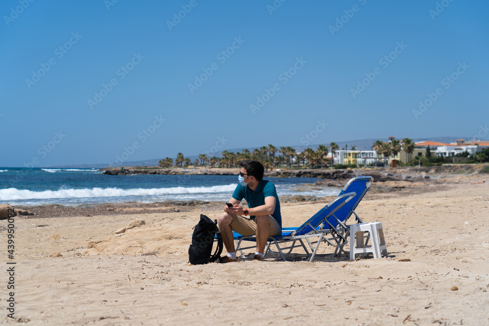 New normal in vacation in time after COVID-19 pandemic. Sad man wearing face mask on beach, sitting on lounge chair and looks out to sea in Cyprus. Male having summer vacation during covid lockdown New normal in vacation in time after COVID-19 pandemic. Sad man wearing face mask on beach, sitting on lounge chair and looks out to sea in Cyprus. Male having summer vacation during covid lockdown