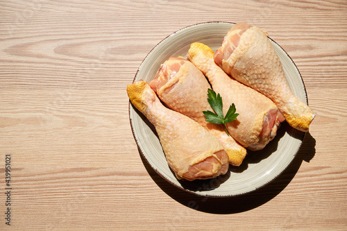 Top view of a raw chicken legs and parsley on a plate on a wooden table next to a window that lets in the light of the sun. Copy space