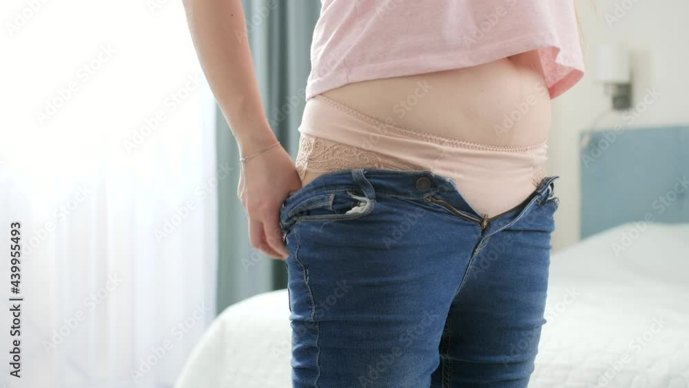 Young woman with wide hips and belly putting on tight jeans and struggling to button them. Concept of excessive weight, obese female, dieting and overweight problems Stock Video | Adobe Stock