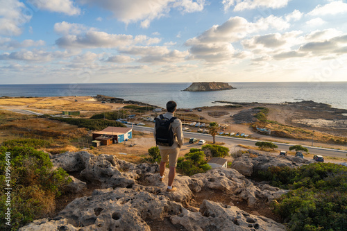 Male tourist with backpack watching beautiful view of Yeronisos Holy Island near coast Agios Georgios Pegeias In Cyprus on sunset. Man hiker looks into distance at deserted island in mediterranean sea