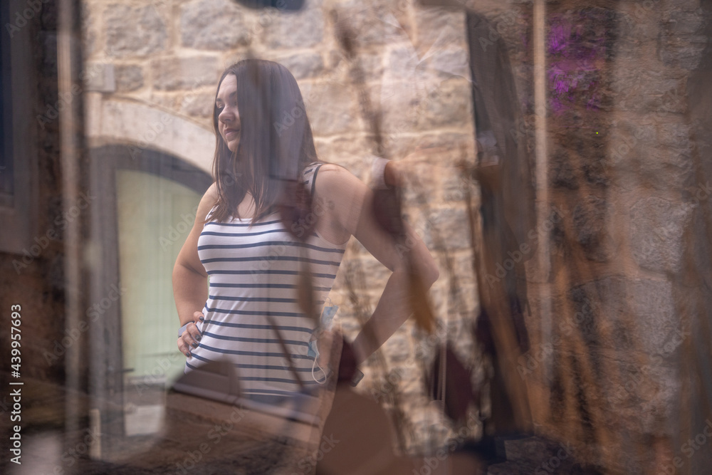 Naklejka premium Young attractive brunette woman is standing in secret courtyard going for a walk, and someone is spying on her from house through the window.