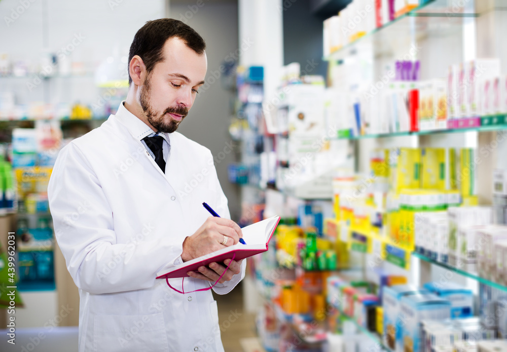 Positive adult man pharmacist writing down assortment of drugs in pharmacy