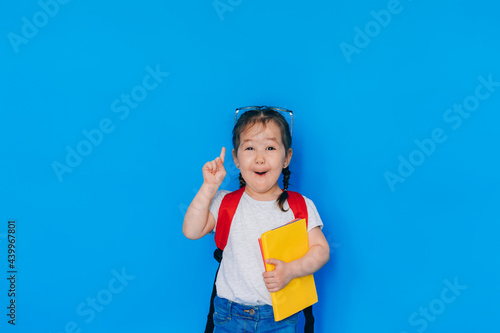 Back to school concept. School girl with red backpack and holding yellow folder in front of blue background. child shows thumb up