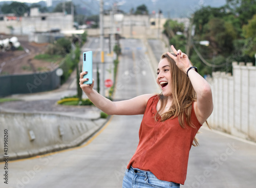 Mujer tomando una foto con el telefono celular, haciendo un gesto de amor y paz a la camara, guatemalteca, rubia, joven y muy bella.