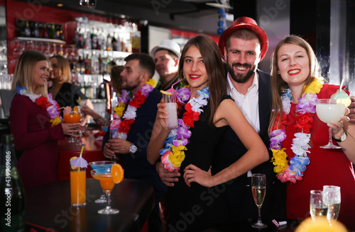 Cheerful guy with two girls dancing and toasting drinks on Hawaiian party in bar