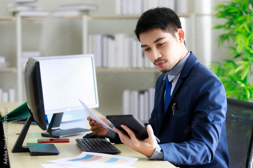 Successful young Asian businessman smiles happily while seeing good news on tablet in office. Business and success concept. Successful young Asian businessman smiles happily while seeing good news on tablet in office. Business and success concept.