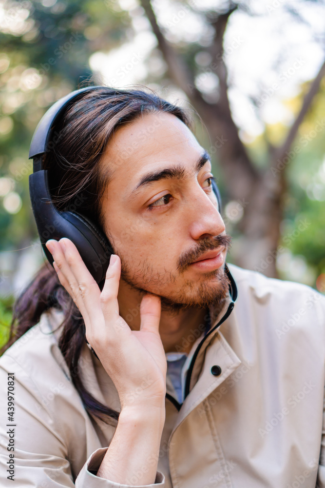 vertical portrait of a long-haired Latino man listening to a podcast with wireless headphones outside in a city park. vertical portrait of a long-haired Latino man listening to a podcast with wireless headphones outside in a city park.