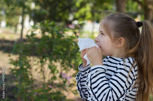 A girl with blond hair in a striped T-shirt sneezes near a bush with flowers in the park, covering her nose with a napkin. Seasonal bloom allergy.