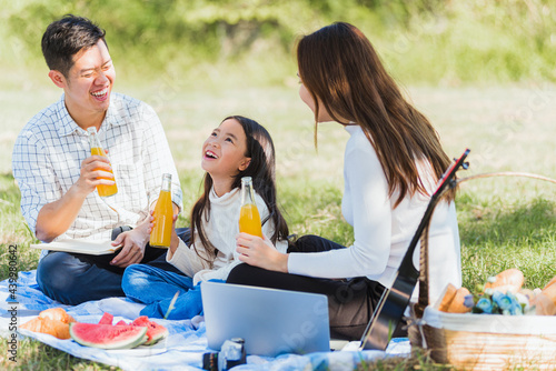 Happy Asian young family father, mother and child little girl having fun and enjoying outdoor sitting on picnic blanket drinking orange juice from glass bottle, Summer resting at a nature garden park photo