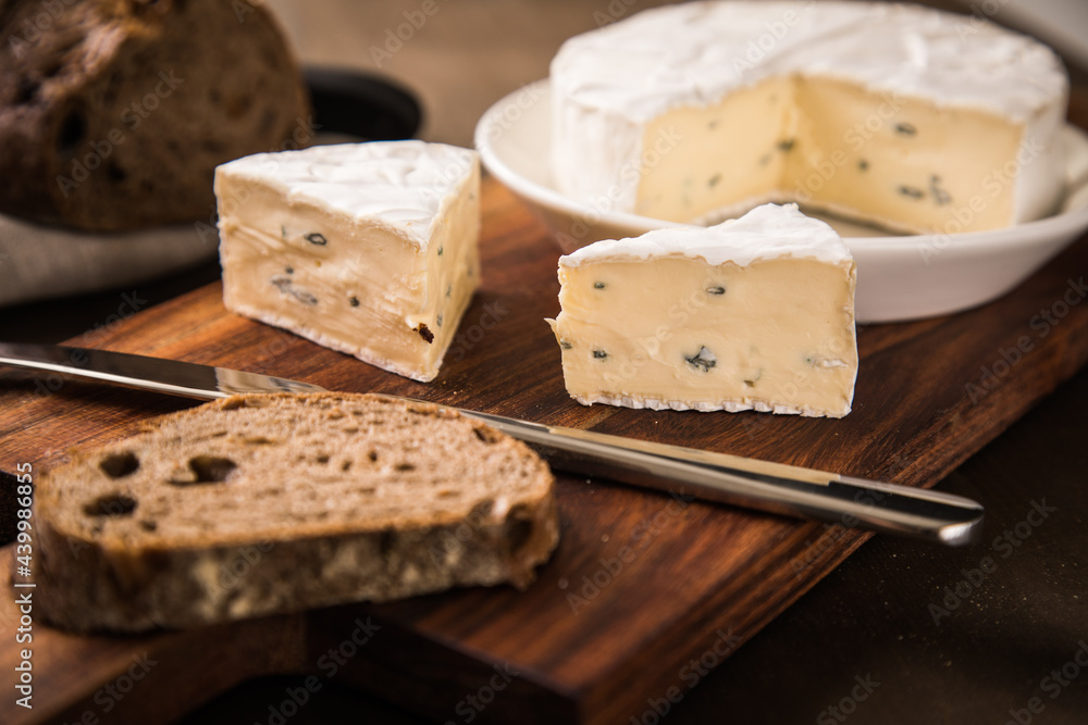 Loaf of soft blue cheese from cow milk on porcelain plate with walnut bread, knife, linen towel and dark brown wooden board as snack or dinner
