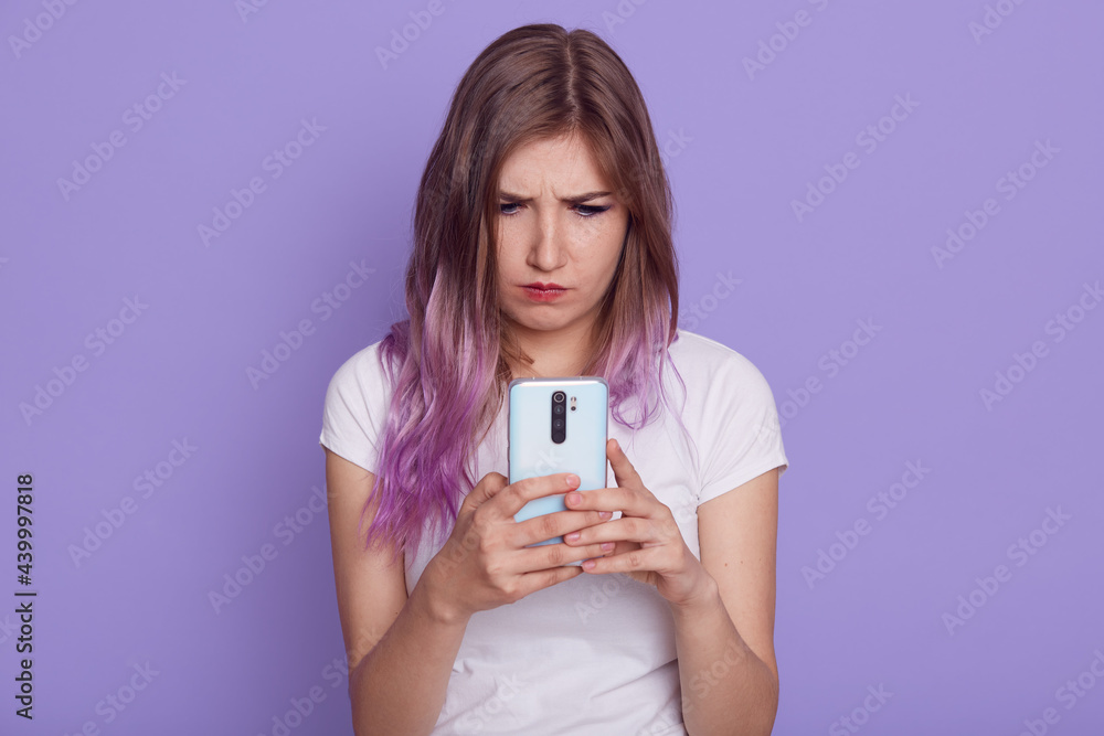 Young beautiful female with bright hair holding cell phone in hands, reading unpleasant news, having upset facial expression, wearing white T-shirt, isolated over lilac background. Young beautiful female with bright hair holding cell phone in hands, reading unpleasant news, having upset facial expression, wearing white T-shirt, isolated over lilac background.