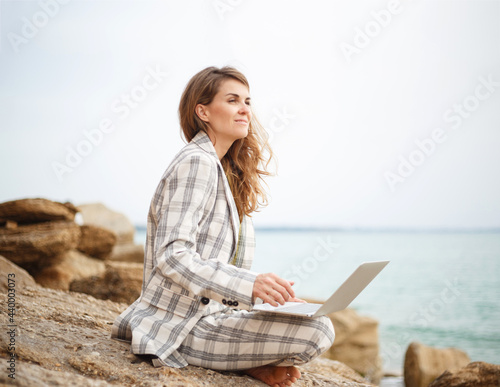 young woman sits on a stone by the sea with a laptop. remote work concept