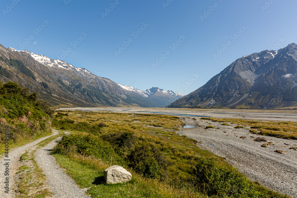 A view of the Hopkins riverbed. Gravel road along the river and the mountain range with snow-capped peaks in the background. Sunny clear summer day. South Island, New Zealand