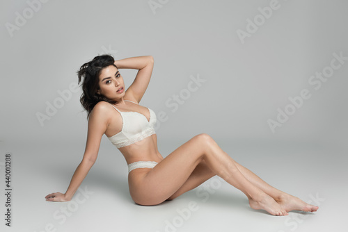 Sensual woman in white lingerie adjusting hair while sitting on grey background