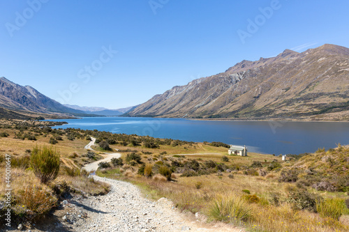 A photo of winding gravel path along North Mavora Lake. Careys Hut and campsite at the lake. Livingstone Mountain range in the background. Sunny summer day. New Zealand landscape.