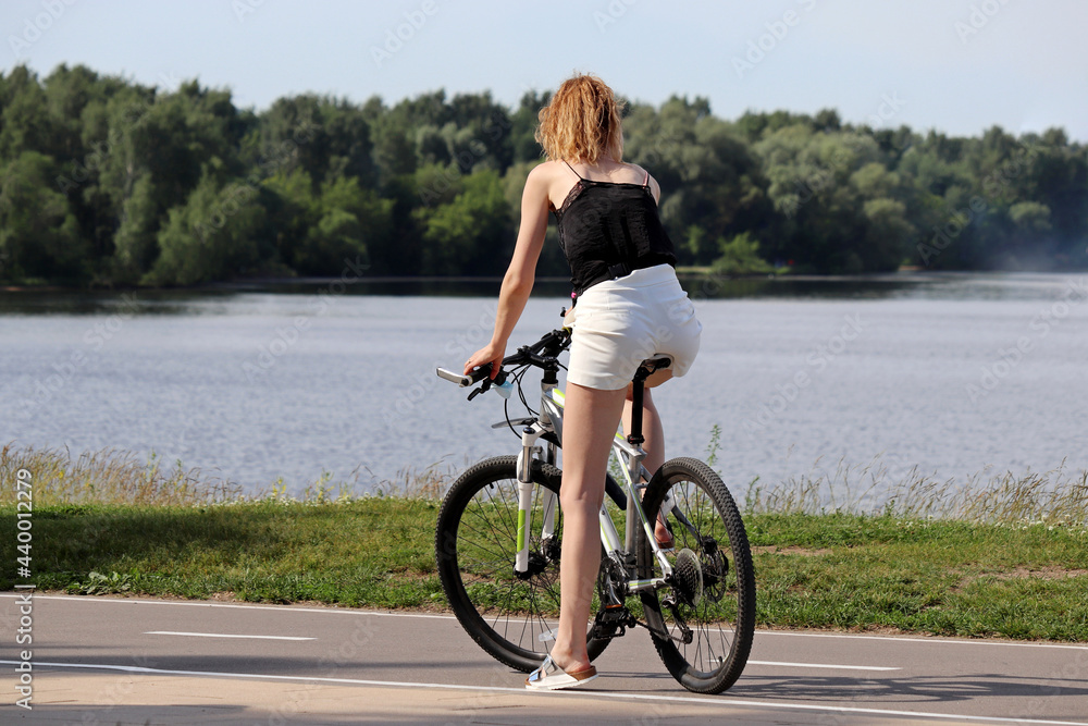 Slim redhead girl in shorts stopped with bicycle on river and forest background. Cycling and summer leisure Slim redhead girl in shorts stopped with bicycle on river and forest background. Cycling and summer leisure