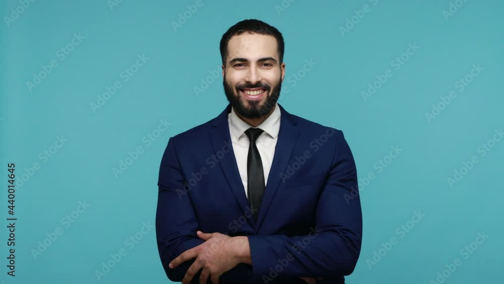 Successful young bearded guy with beautiful smile turns to camera and folds his arms, has positive facial expression, rejoices in good business deal. Indoor studio shot isolated on blue background. Successful young bearded guy with beautiful smile turns to camera and folds his arms, has positive facial expression, rejoices in good business deal. Indoor studio shot isolated on blue background.