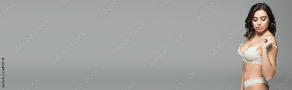 Seductive woman looking at shoulder while taking off bra isolated on grey, banner Seductive woman looking at shoulder while taking off bra isolated on grey, banner