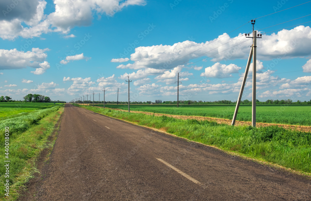 Village Road with power line, green fields and blue sky with clouds