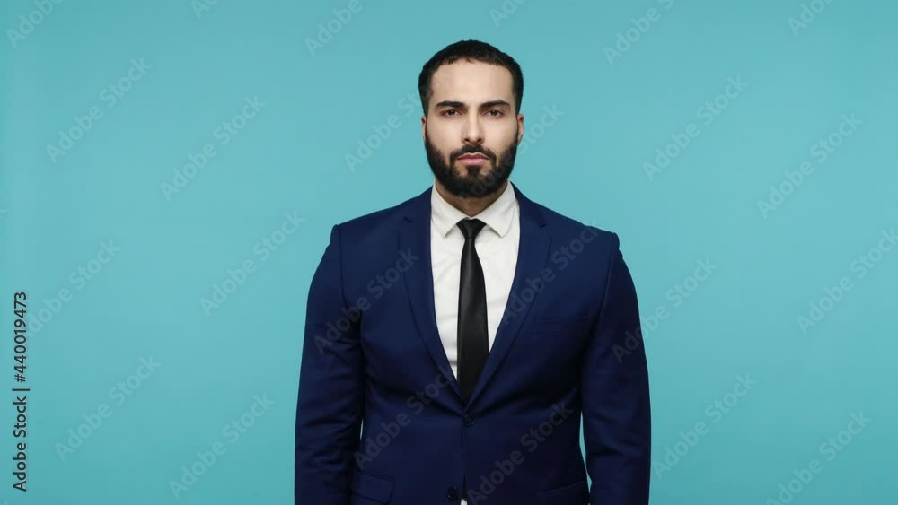 Bearded man in official style suit saluting with hand, saying yes sir and looking at camera with positive expression, pretending to listen order. Indoor studio shot isolated on blue background. Bearded man in official style suit saluting with hand, saying yes sir and looking at camera with positive expression, pretending to listen order. Indoor studio shot isolated on blue background.
