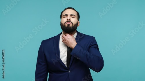 Unhappy frustrated man with beard suffers from throat pain, touches neck with hands, looks dissatisfied at camera, wears black formal suit. Indoor studio shot isolated on blue background.