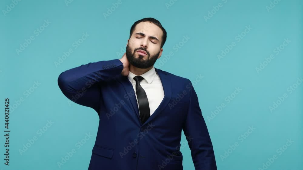 Displeased young man with beard feels strong pain in neck, tilts head smirks face dressed in black formal suit, has problems with spine. Indoor studio shot isolated on blue background. Displeased young man with beard feels strong pain in neck, tilts head smirks face dressed in black formal suit, has problems with spine. Indoor studio shot isolated on blue background.