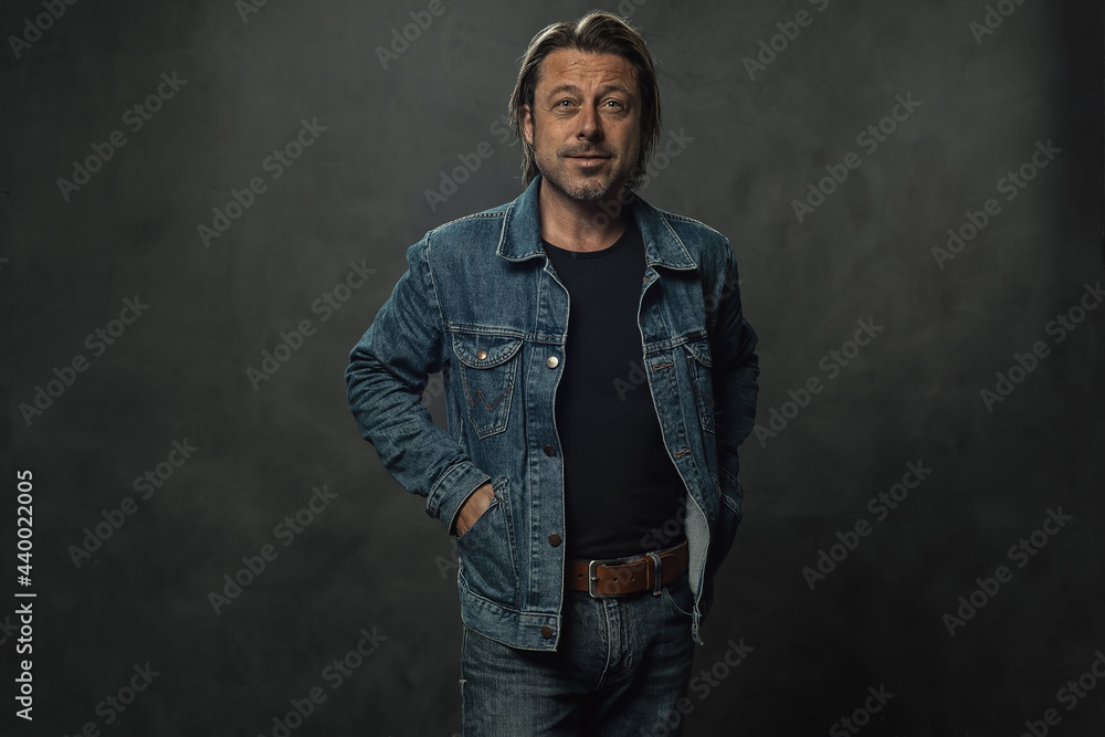 Obraz premium Blond man in jeans with a brown leather belt and a denim jacket in front of a dark grey wall.