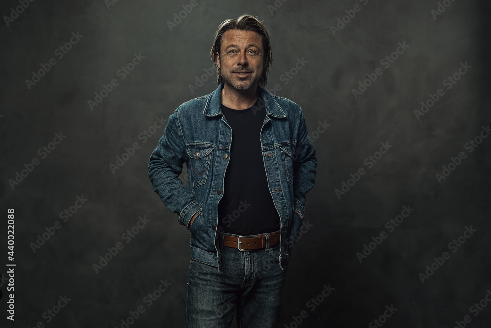Blond man in jeans with a brown leather belt and a denim jacket in front of a dark grey wall. Blond man in jeans with a brown leather belt and a denim jacket in front of a dark grey wall.