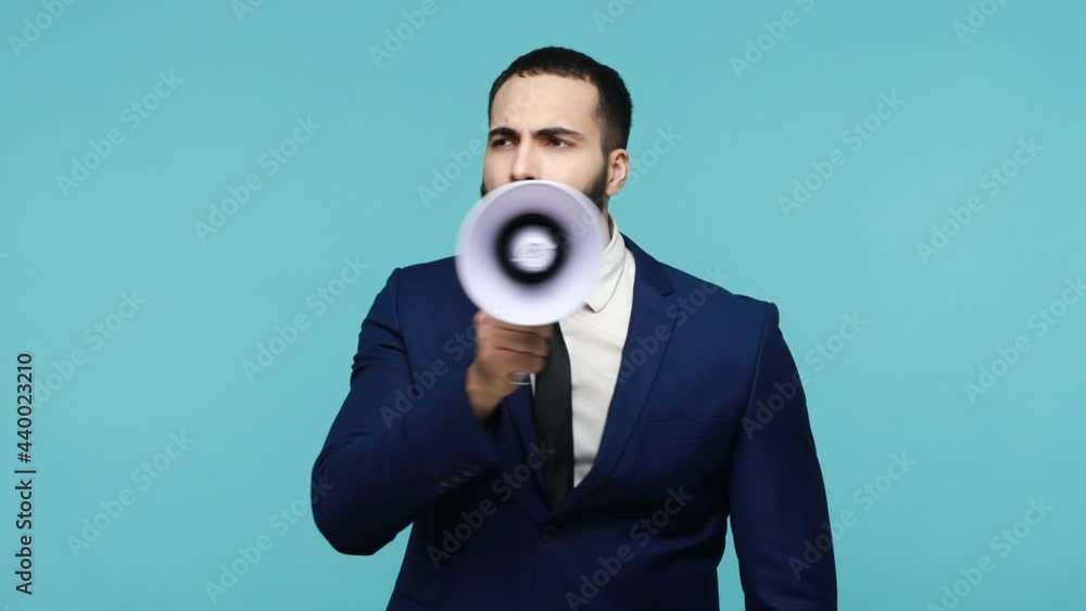 Serious young brunet man speaker talking holding megaphone near mouth, trying to explain, expressing his point of view, making announcement. Indoor studio shot isolated on blue background. Serious young brunet man speaker talking holding megaphone near mouth, trying to explain, expressing his point of view, making announcement. Indoor studio shot isolated on blue background.