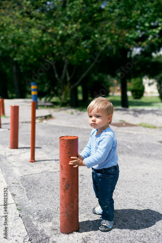 Kid stands on the street near a little red metal post against a background of trees © Nadtochiy