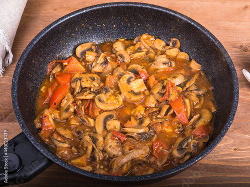 Cooking homemade champignon mushroom sauce and vegetables, tomatoes and onions in a pan. Frying pan with ready-made stewed chopped mushrooms with red tomato and onion sauce on the table, close-up