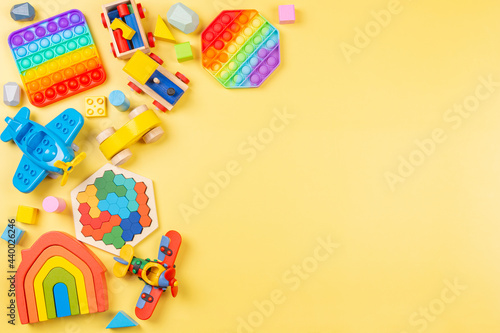 Baby kids toys background with wooden rainbow color house, train, car, plane, pop it fidget toys and colorful blocks on yellow background. Top view, flat lay photo