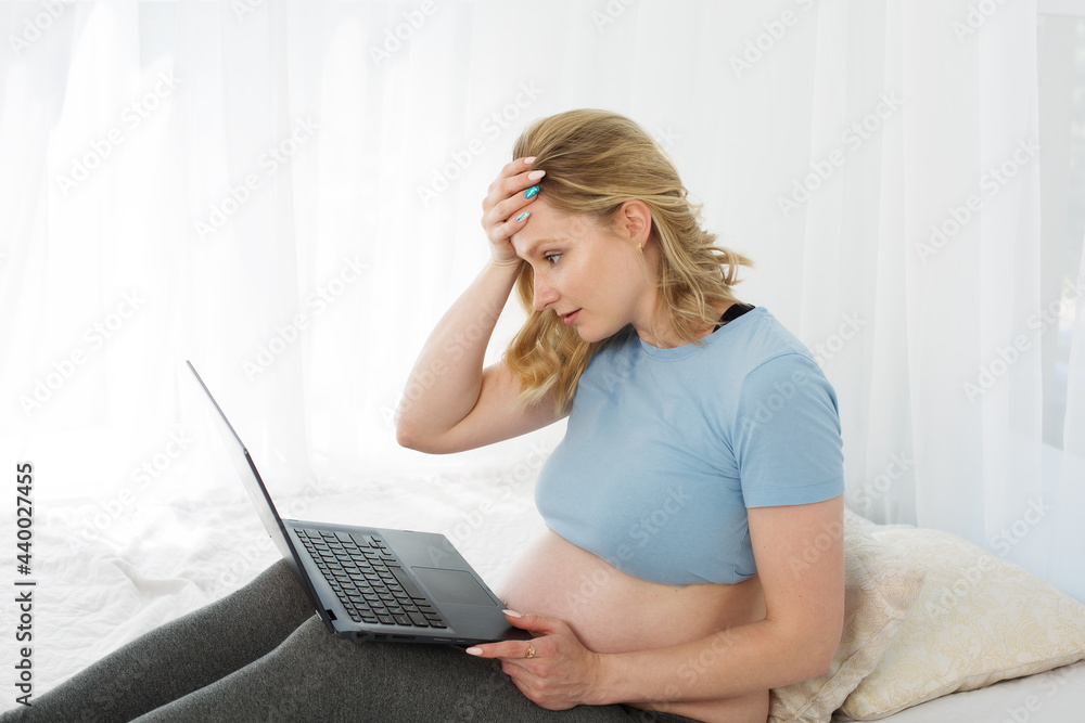 Fototapeta premium Blonde pregnant woman working on a computer, surprised, upset, holding her head with her hand online shopping