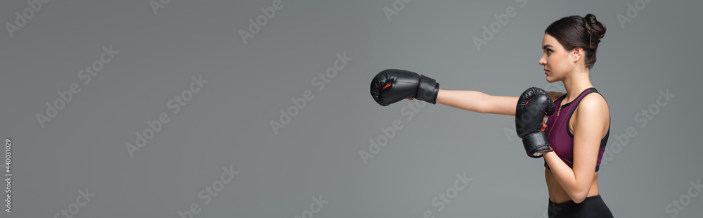 side view of sportive woman training in boxing gloves isolated on grey, banner.