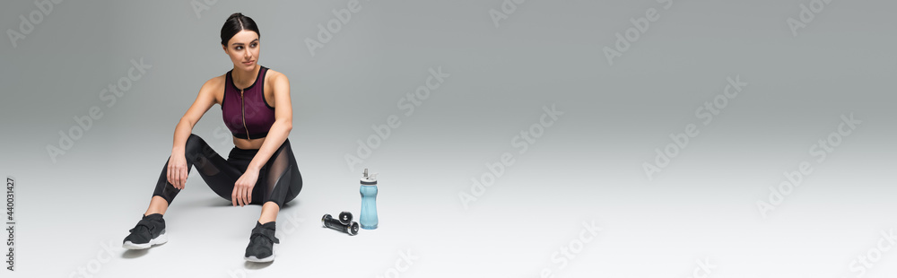 Fototapeta premium sportive woman looking away while sitting near sports bottle and dumbbells on grey background, banner.