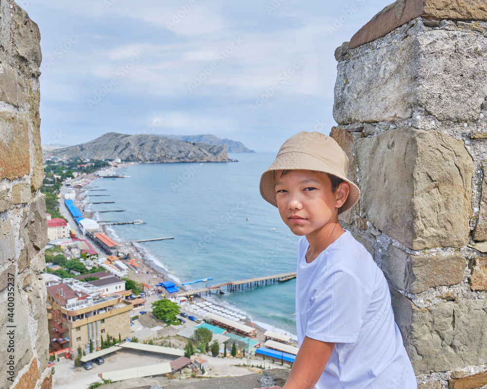 Asian boy l near the wall of ancient fortress against background of embankment of town of Sudak, Crimean peninsula.