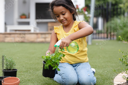 Smiling hispanic girl gardening, kneeling and watering plant in pot photo