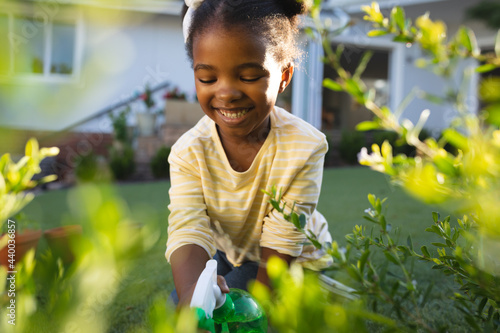 Smiling african american girl gardening, kneeling and watering plants in sunny garden