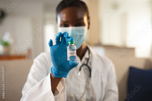 African american female doctor in face mask and gloves holding vial of covid 19 vaccine photo