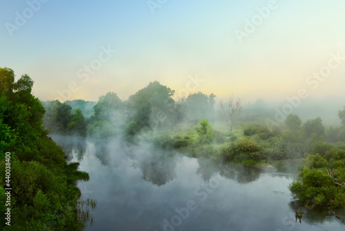 small quiet river at dawn, green overgrown coast meandering river, fog, mist over the water. Summer morning 