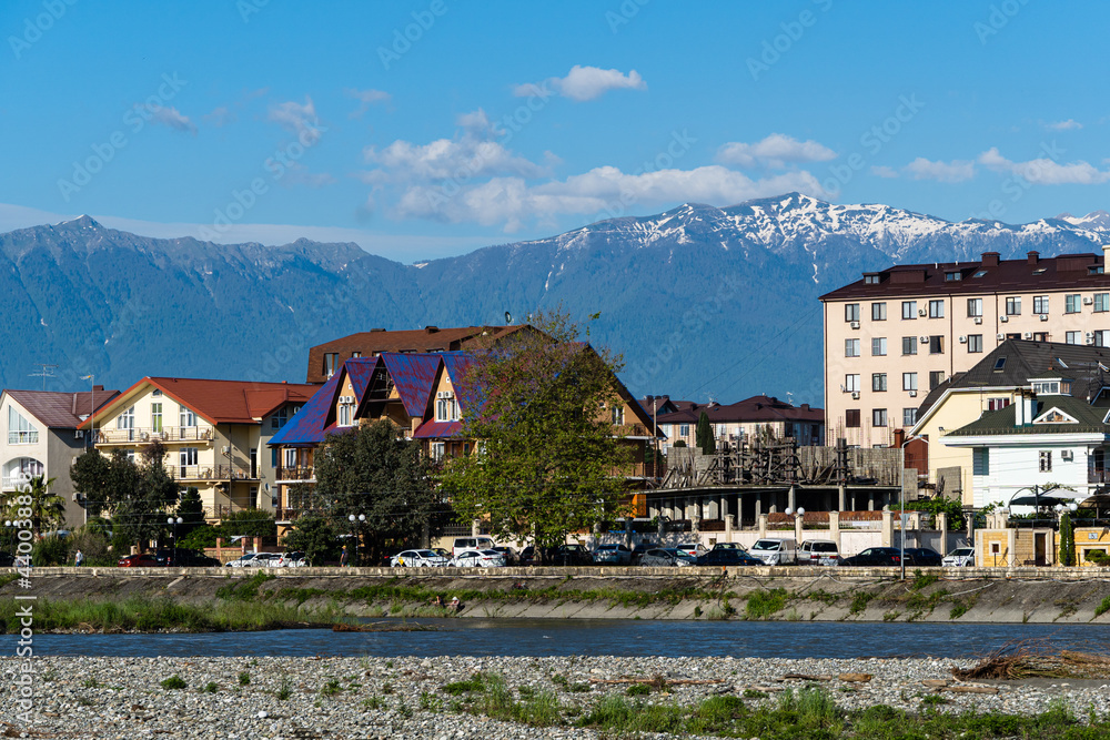 Residential buildings in federal territory of Sirius against backdrop of snow-capped peaks. Caucasus mountains. Imeretinskaya lowland. In foreground is Mzymta.Adler. Sochi, Russia - May 18, 2021