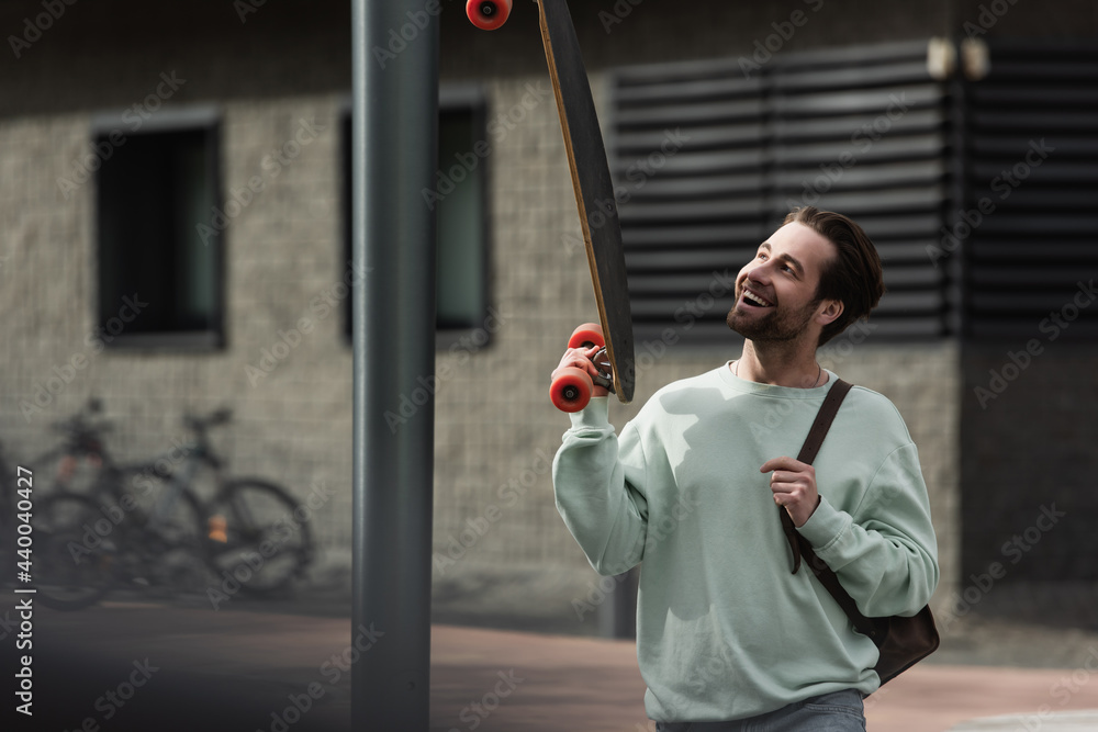 Fototapeta premium cheerful bearded man in sweatshirt holding longboard and leather strap of backpack outside.