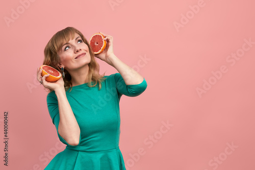Young woman with halves of a grapefruit in her hands holds an ear to her ears, a cheerful girl in a green dress with grapefruits on a pink background, copy space